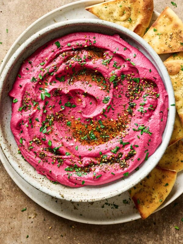 vibrant pink beet hummus in a ceramic bowl, topped with parsley and za'atar, on a plate with pita bread.