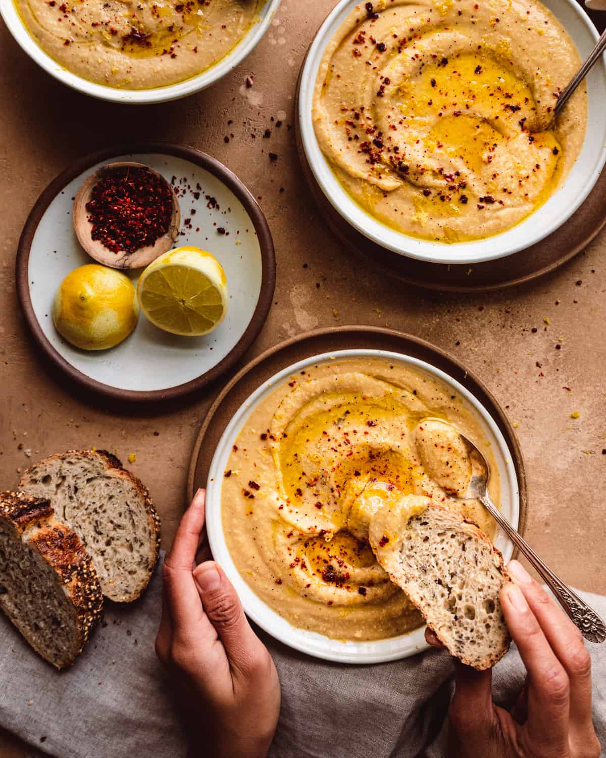 woman's hands dunking bread into a bowl of creamy vegan cauliflower soup on a brown table with bread and lemons on the side.