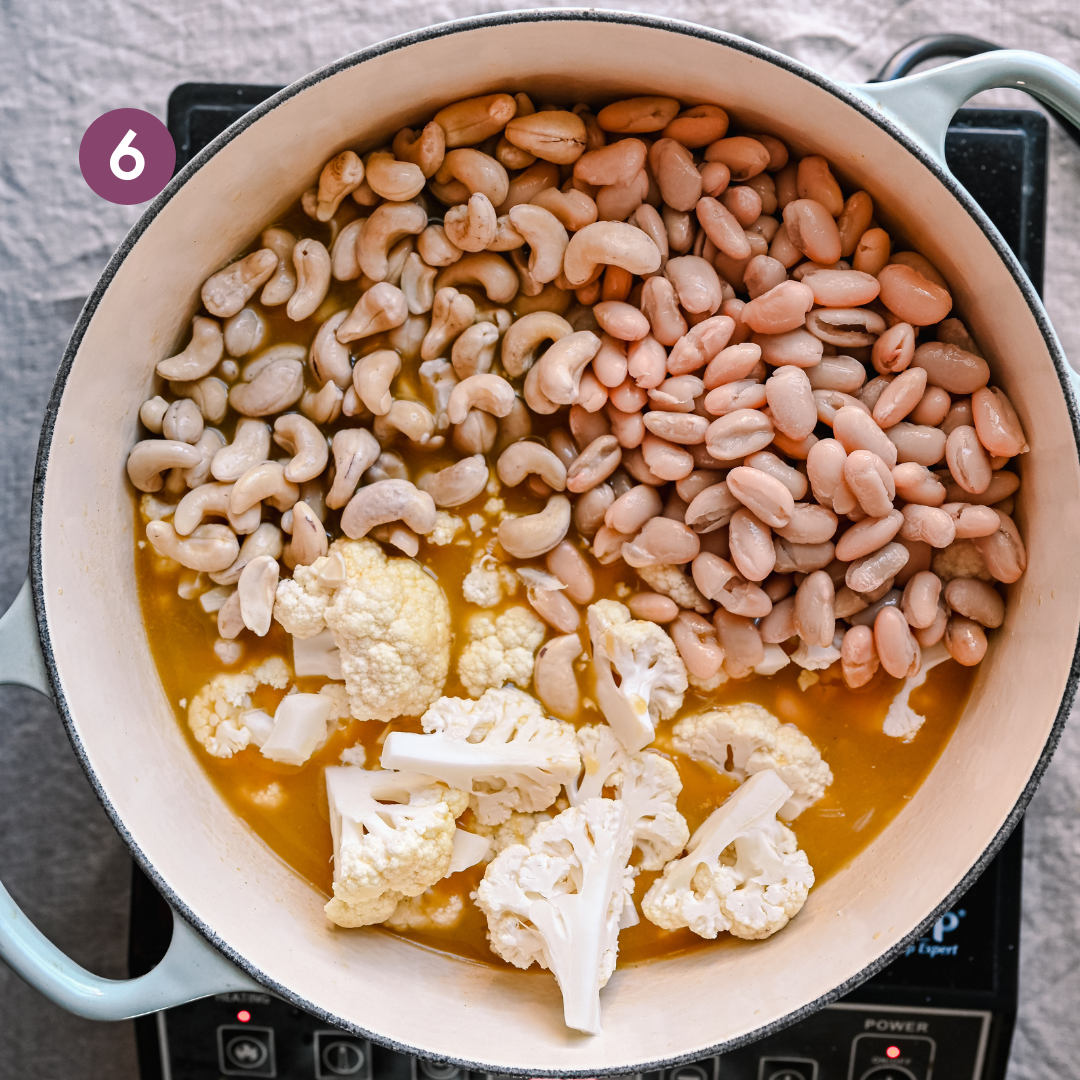 cauliflower florets, cannellini beans, and raw cashews sitting on top of vegetable broth for a soup in a dutch oven.