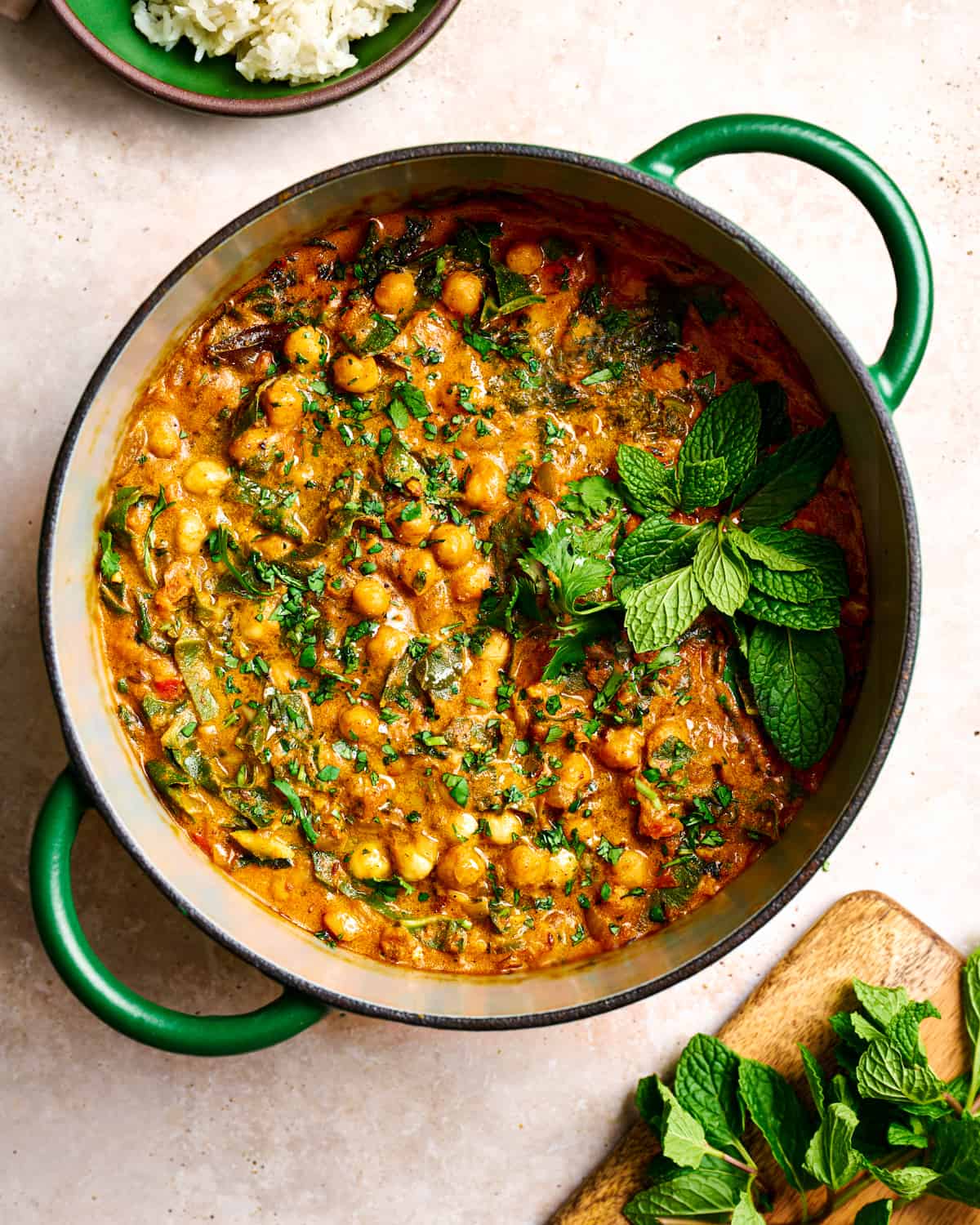 vegan chickpea curry in a green dutch oven garnished with mint and cilantro on a light pink surface.