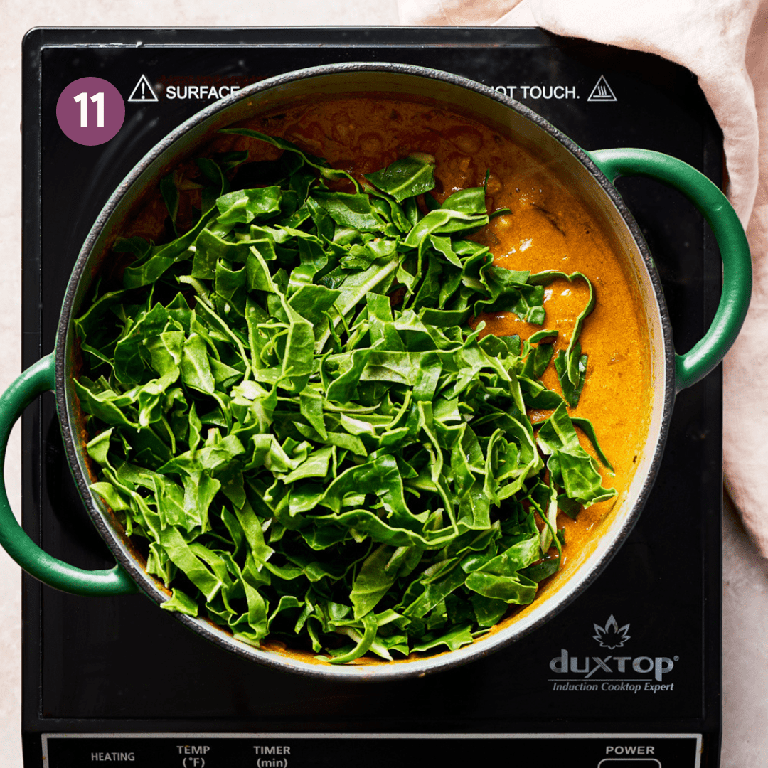 sliced leafy greens sitting on top of a orange-colored vegan curry in a green dutch oven.