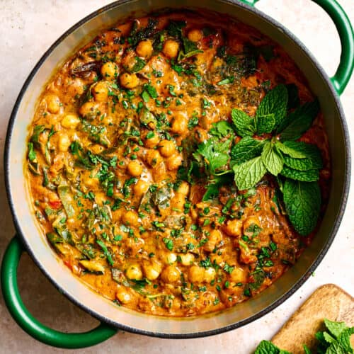 vegan chickpea curry in a green dutch oven garnished with mint and cilantro on a light pink surface.