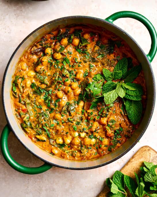 vegan chickpea curry in a green dutch oven garnished with mint and cilantro on a light pink surface.