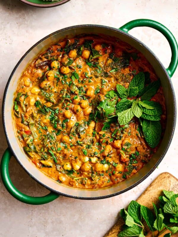 vegan chickpea curry in a green dutch oven garnished with mint and cilantro on a light pink surface.