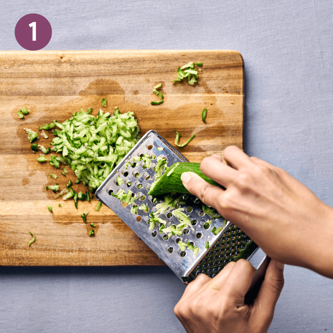 Cucumber being grated onto a wood cutting board.