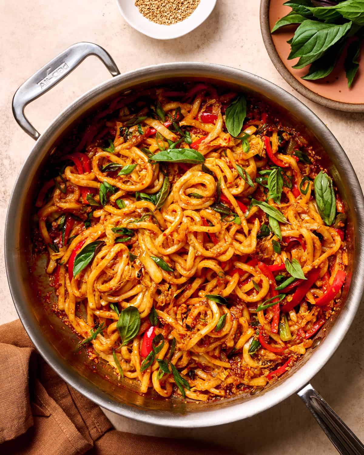 Finished gochujang noodles in a saucepan and small ingredient bowls on a tan table.