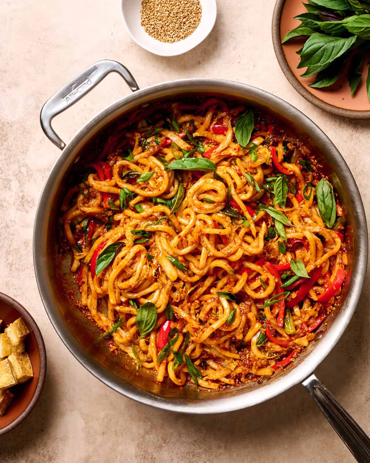 Finished gochujang noodles in a saucepan and small ingredient bowls on a tan table.