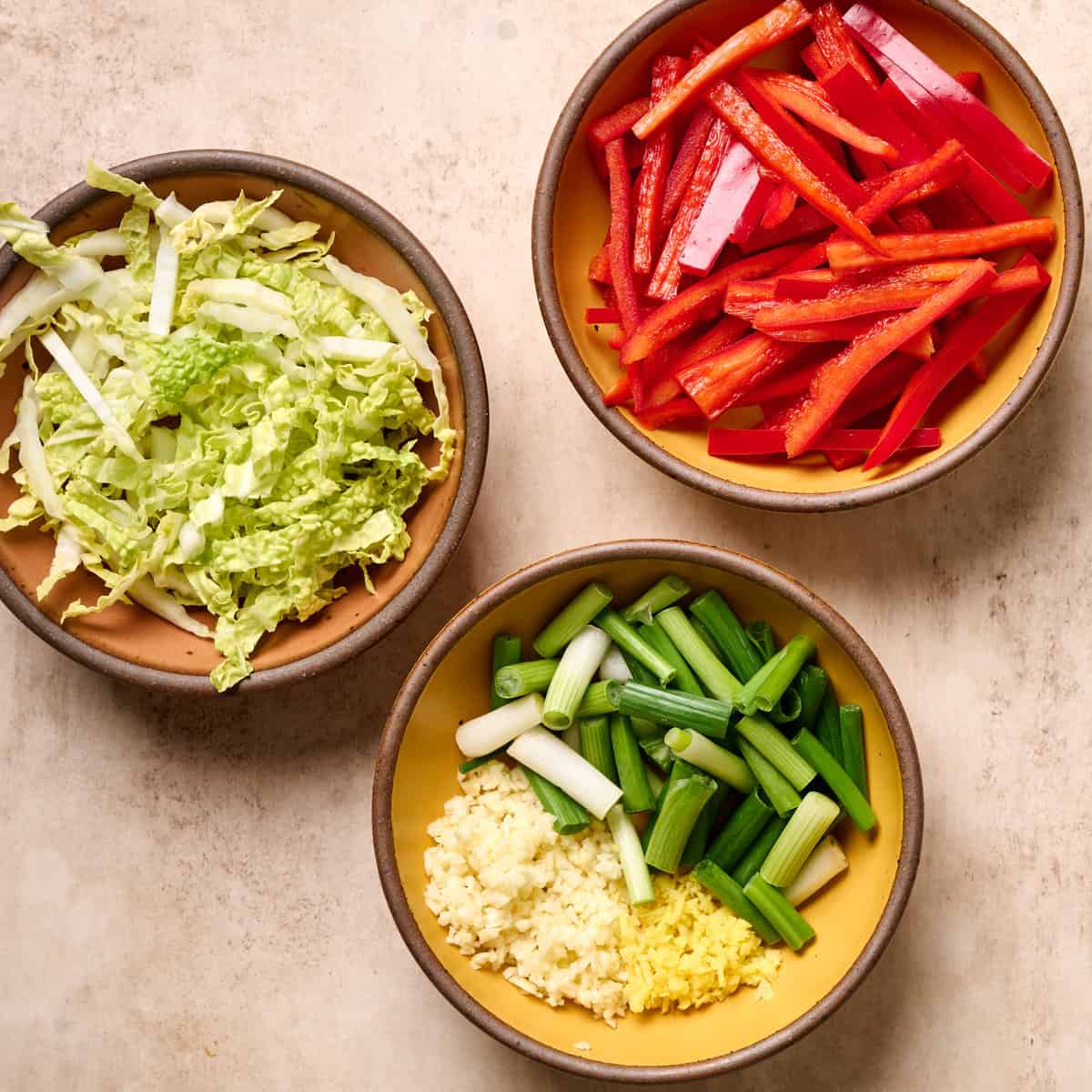 Sliced vegetables and aromatics in three small bowls on a tan table.