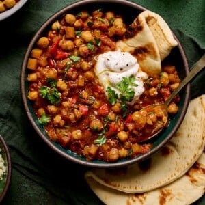 bowl of vegan tagine with chickpeas topped with yogurt and pita on a green table with green glasses.