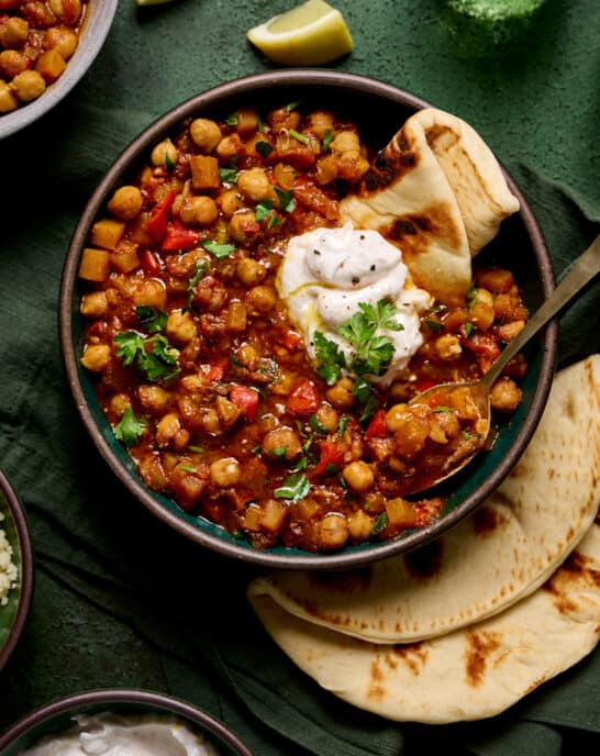 bowl of vegan tagine with chickpeas topped with yogurt and pita on a green table with green glasses.