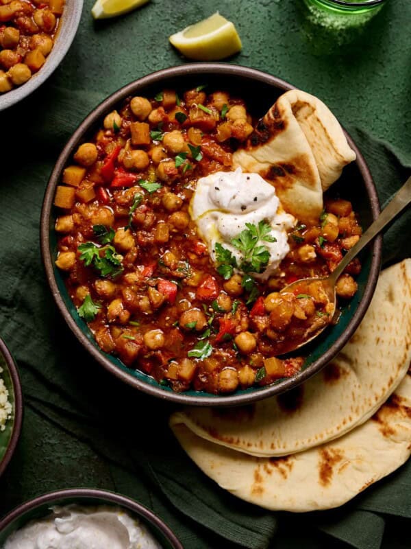 bowl of vegan tagine with chickpeas topped with yogurt and pita on a green table with green glasses.