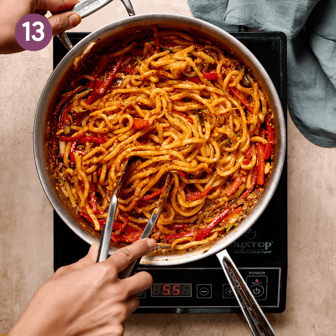 Person using tongs to stir udon noodles into saucy veggie mixture.