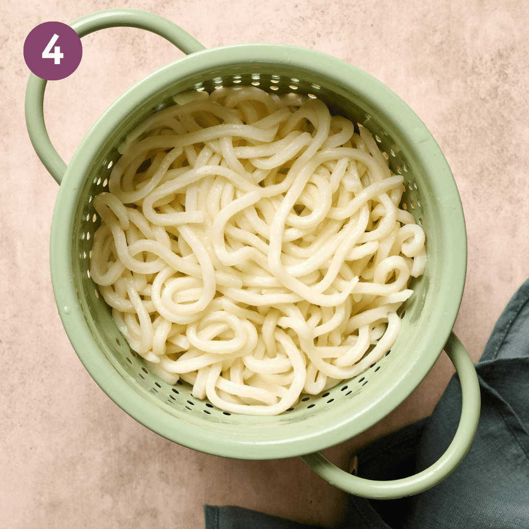 Cooked udon noodles in a green colander on a table.