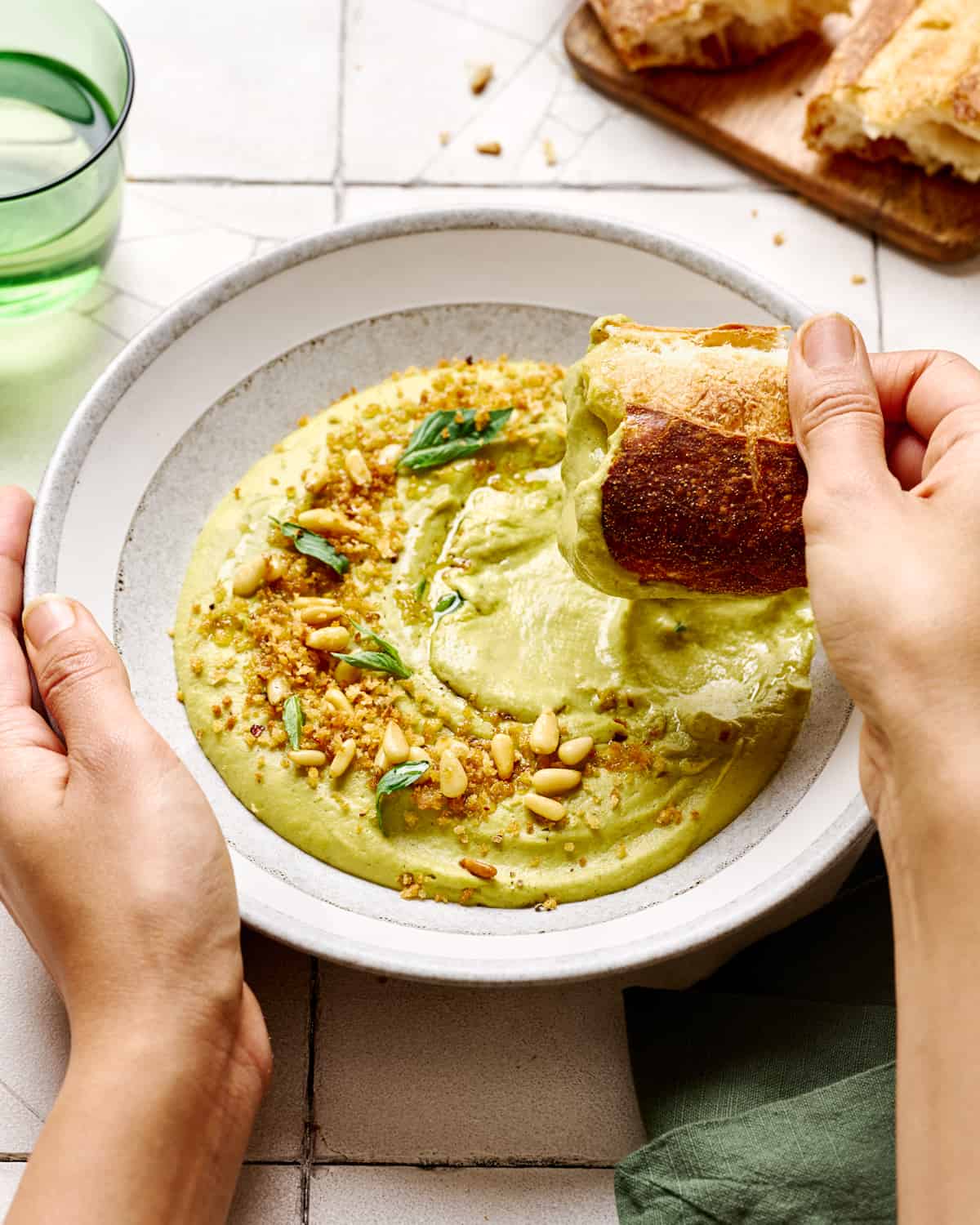 Person holding bowl while dipping chunk of bread into broccoli soup in white bowl.