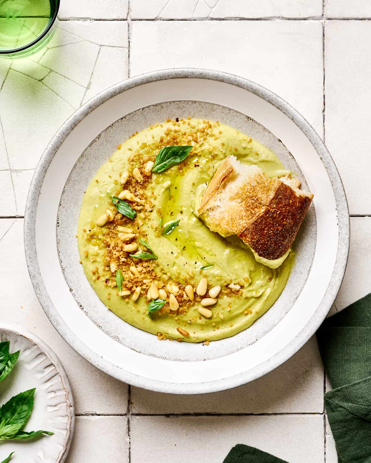 Chunk of bread into broccoli soup in white bowls on white tile table.