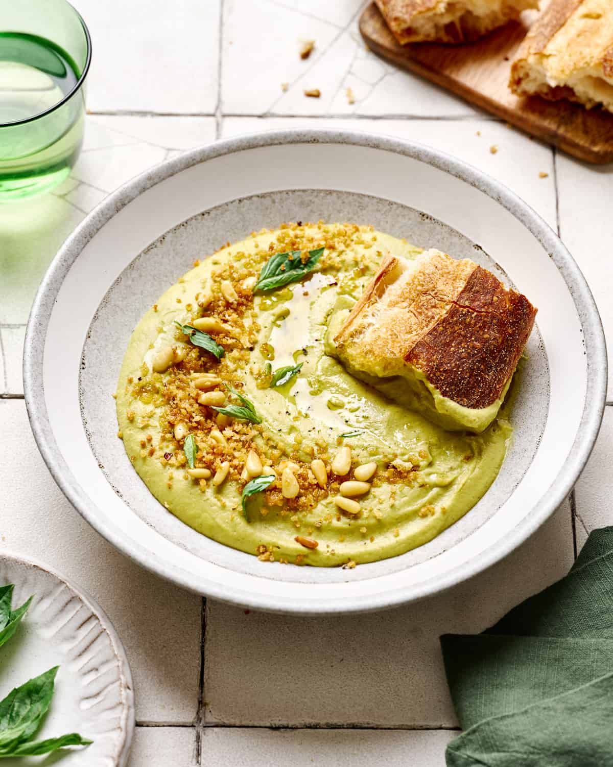 Chunk of bread into broccoli soup in white bowls on white tile table.