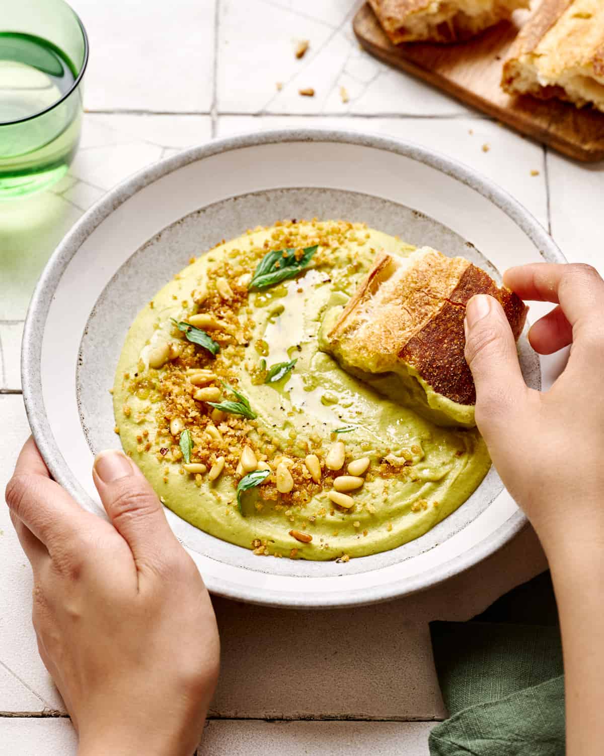 Person dipping chunk of bread into broccoli soup in white bowl.