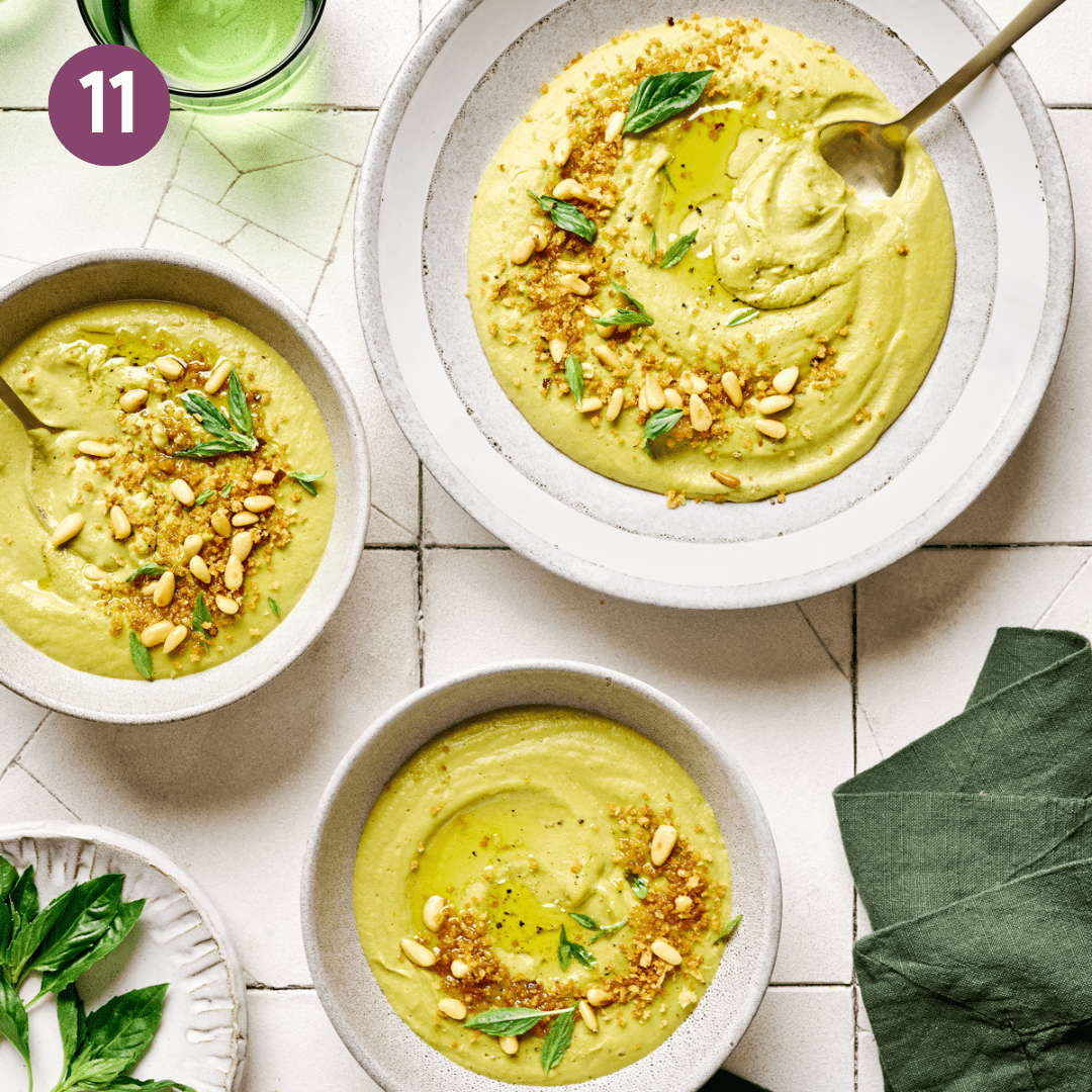 Broccoli soup topped with nutty crumbles in three white bowls on white tile table.