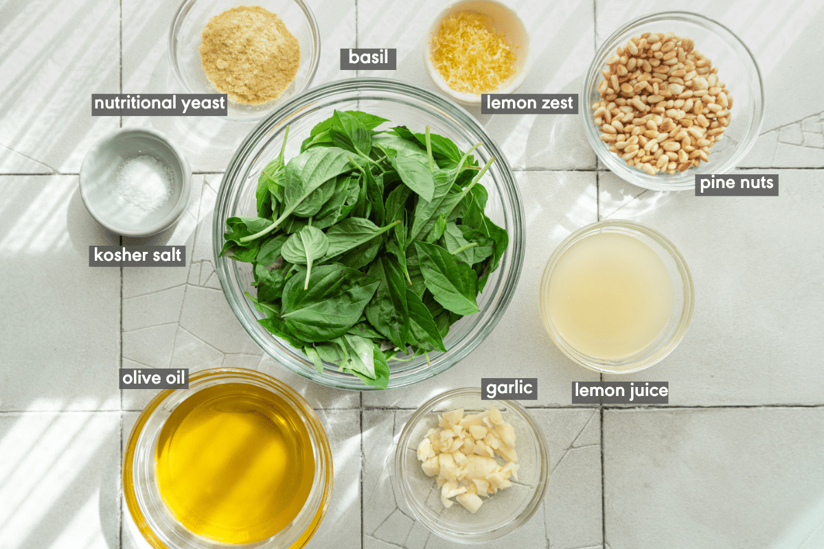Basil pesto ingredients in various glass bowls on a white tile table.
