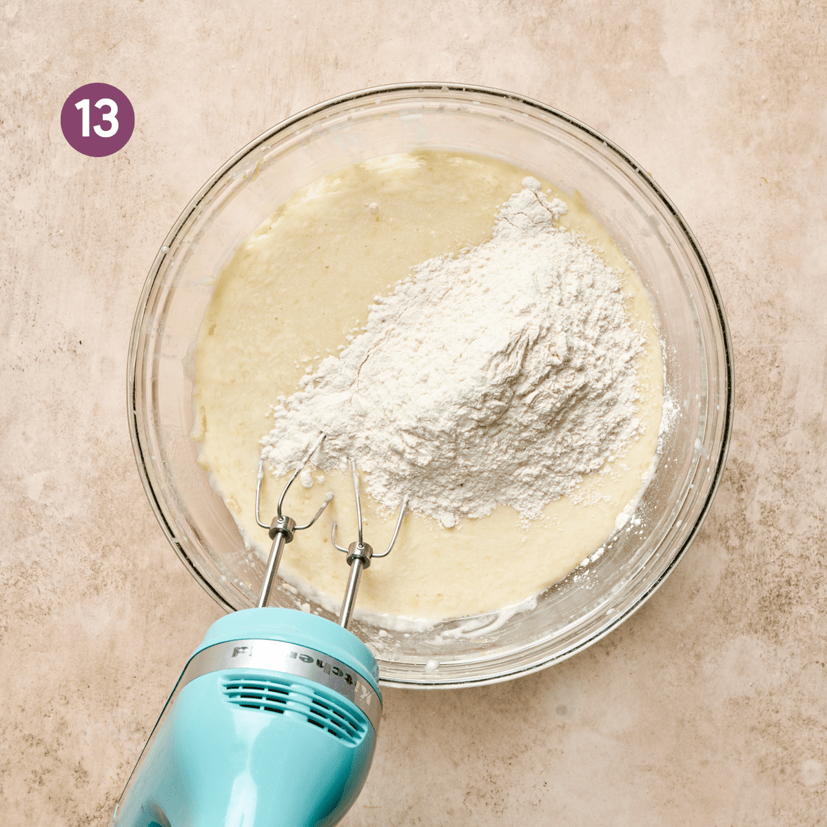 light blue electric mixer in a glass bowl of liquid ingredients with flour on top.