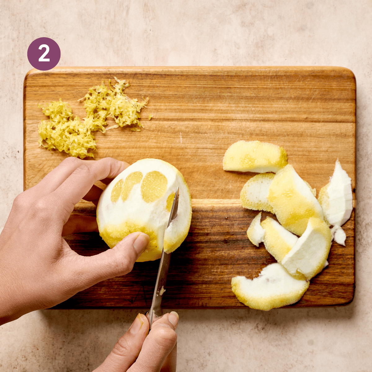woman's hands using a knife to peel a zested lemon on a wooden cutting board.