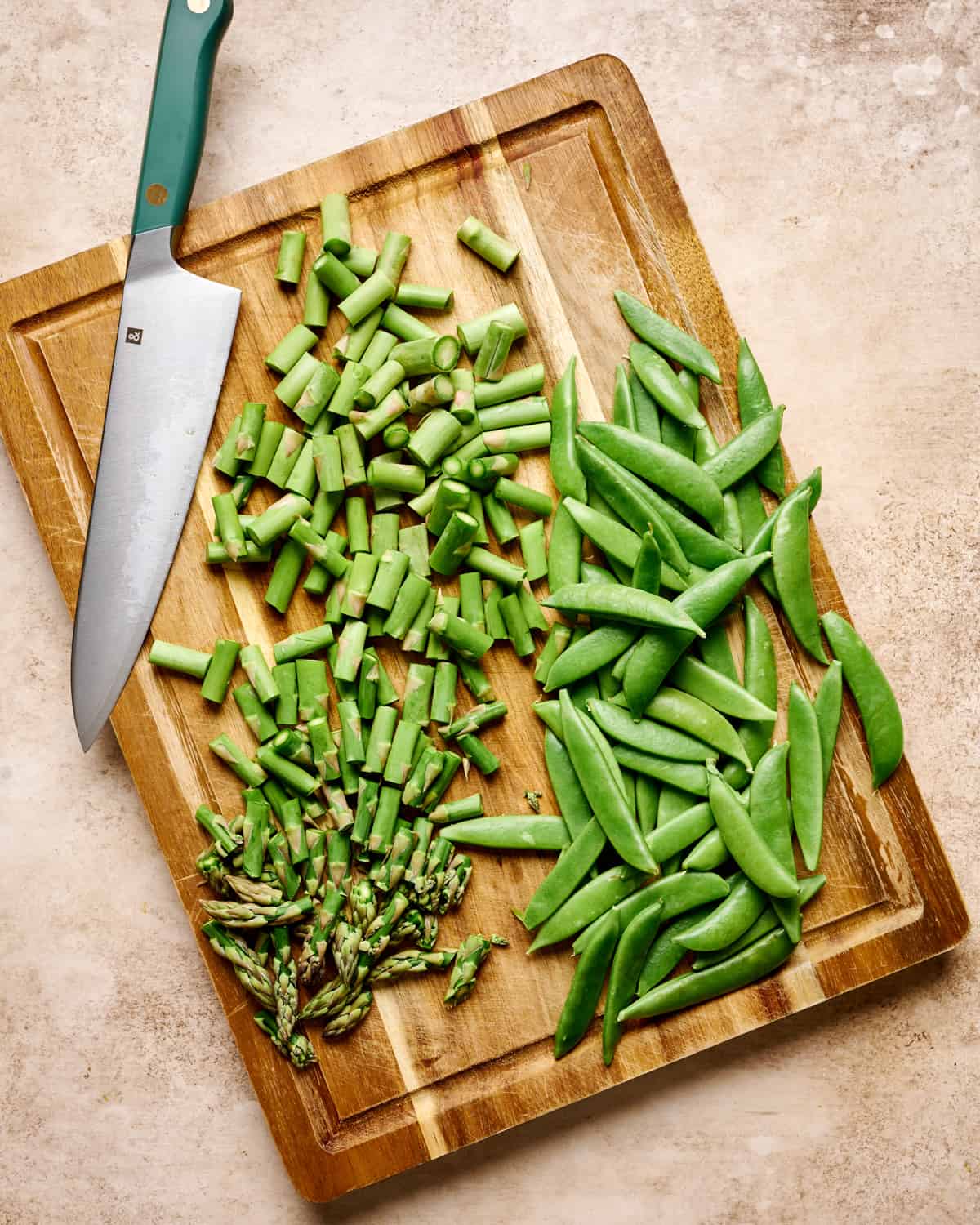 A knife, chopped asparagus and snap peas on a wooden cutting board.
