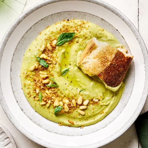 Hunk of sourdough in a bowl of creamy vegan broccoli soup in a white bowl on white tile table.