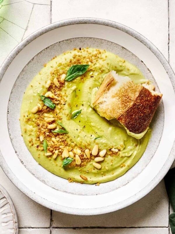 Hunk of sourdough in a bowl of creamy vegan broccoli soup in a white bowl on white tile table.