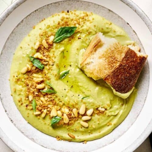 Hunk of sourdough in a bowl of creamy vegan broccoli soup in a white bowl on white tile table.