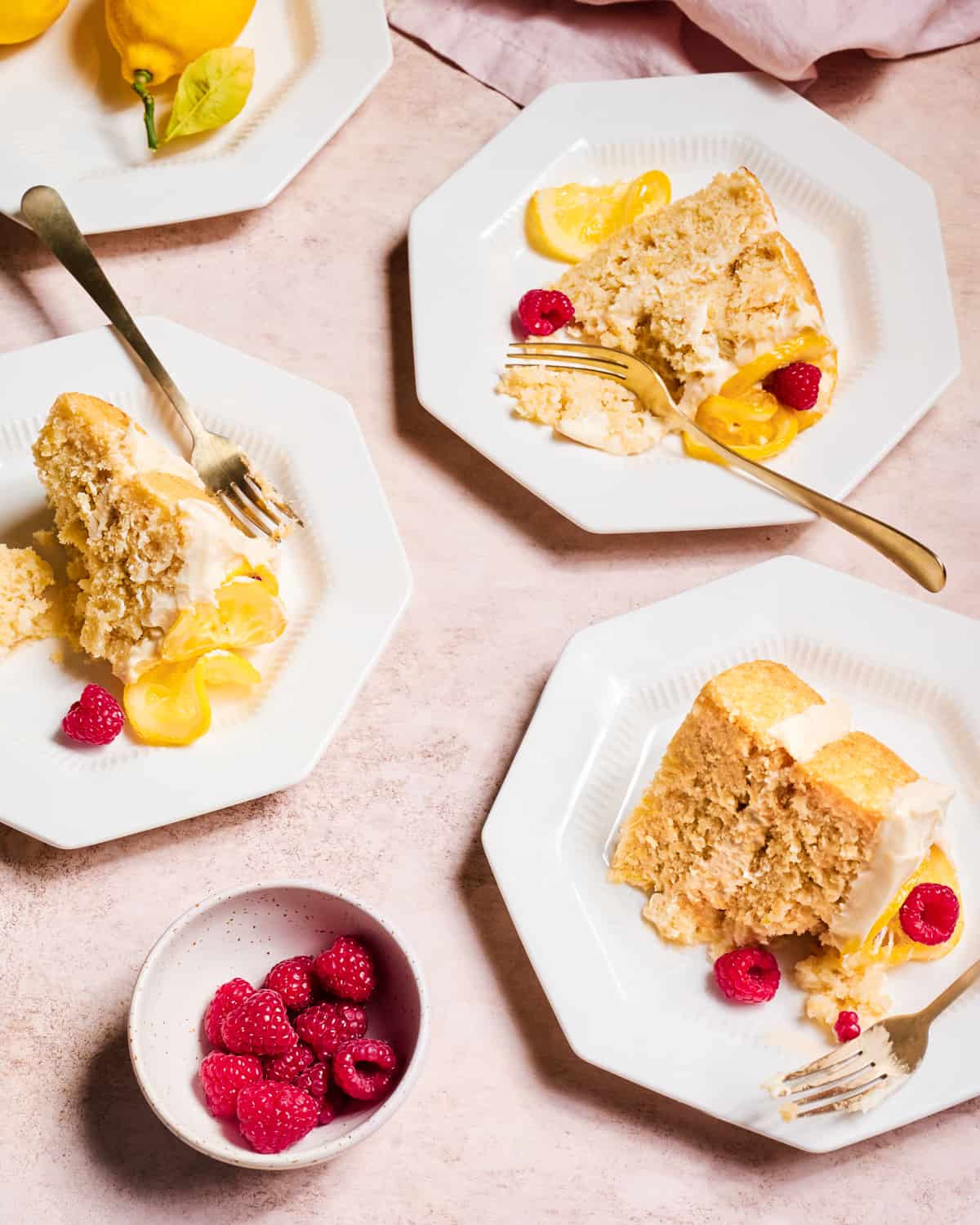 three plates with slices of vegan lemon cake with raspberries and candied lemons on pink surface.