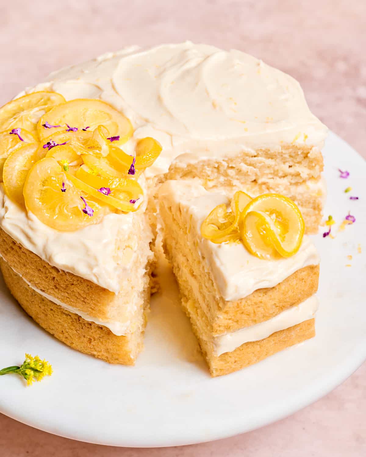 vegan lemon cake with cream cheese frosting on a white cake stand, with one slice out.