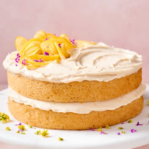 two-layer vegan lemon cake with lemon cream cheese frosting on a white cake stand topped with candied lemons.