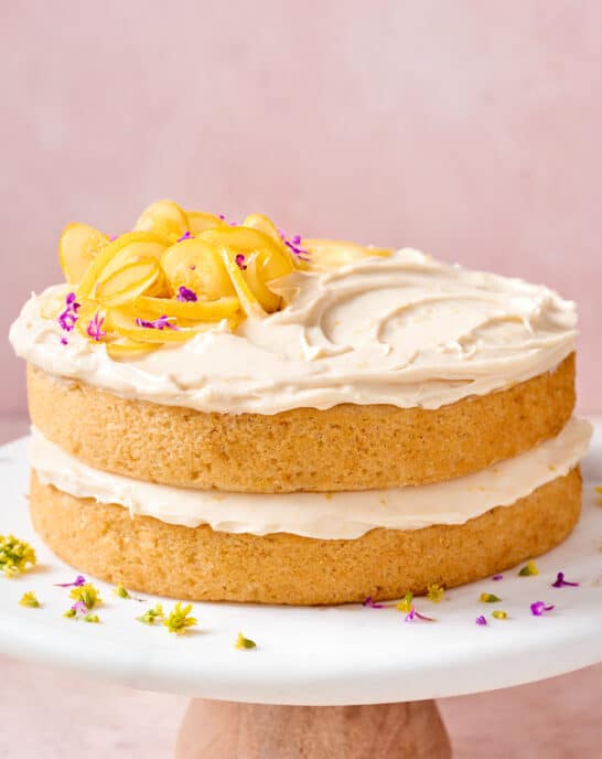 two-layer vegan lemon cake with lemon cream cheese frosting on a white cake stand topped with candied lemons.