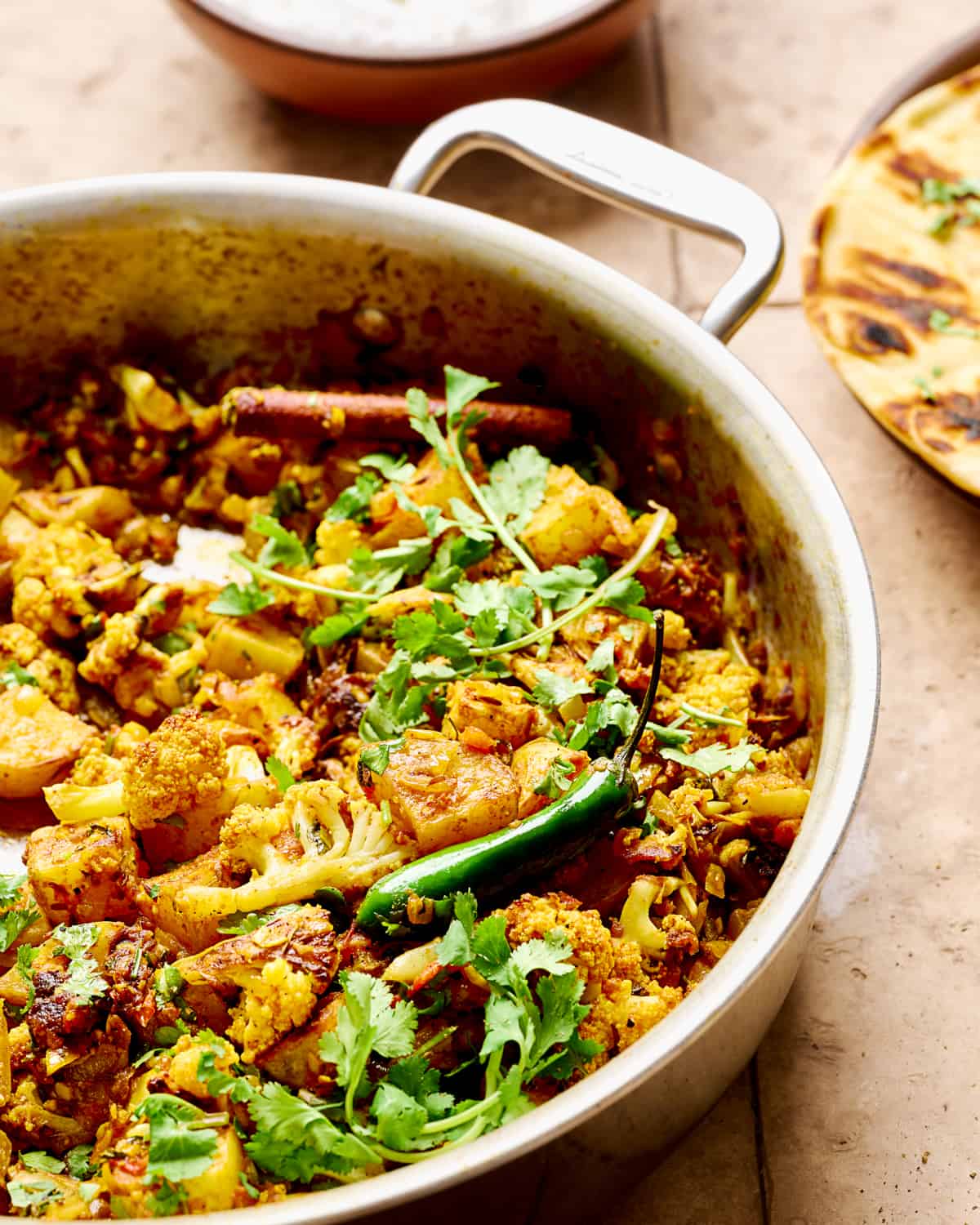 Side view of aloo gobi in a deep sauté pan on a brown table.