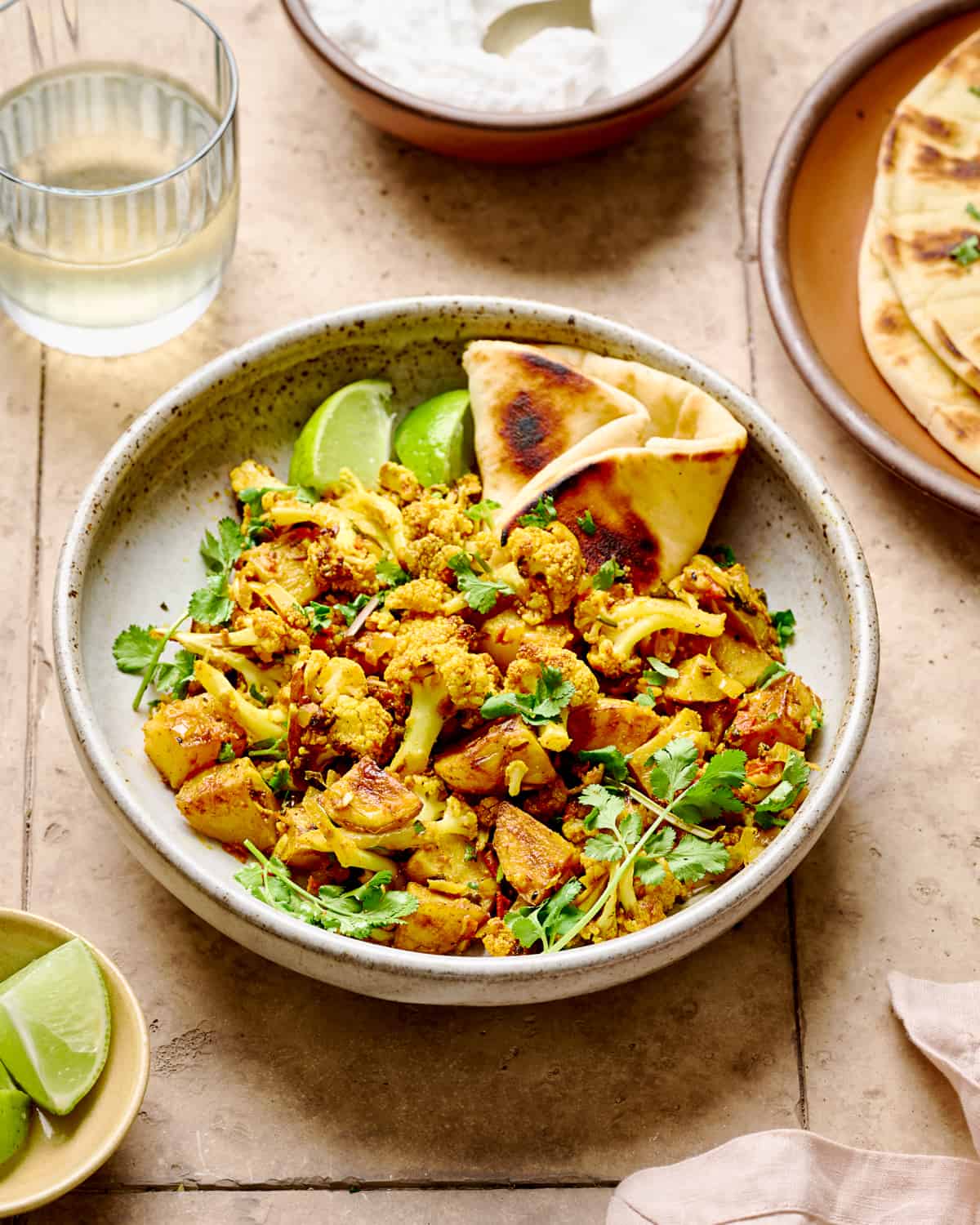 Aloo Gobi, naan and lime wedges in a white bowl on a brown tile table.