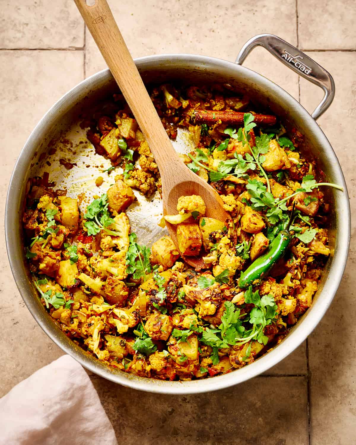 Overhead view of a spoon in pan of aloo gobi on a brown table.