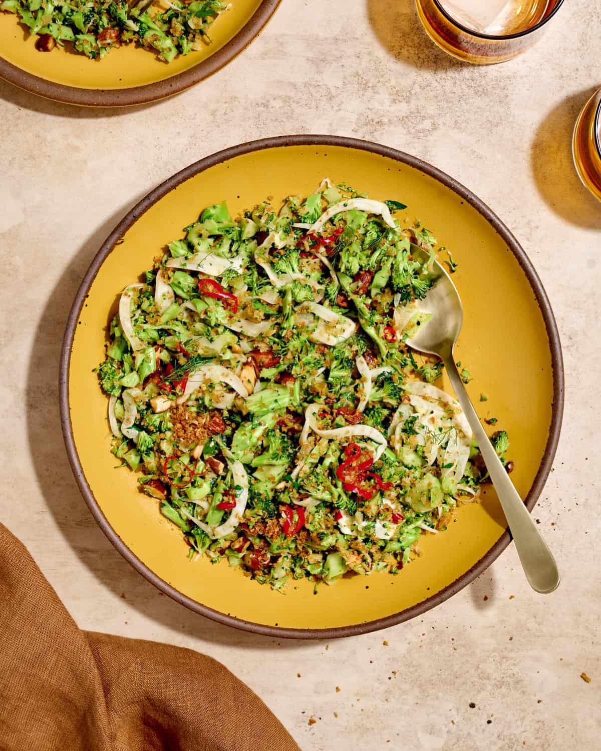 Overhead view of broccoli salad and a serving spoon on a yellow plate on a table.