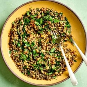 Spoon and fork in lentil salad in a yellow bowl on green table.