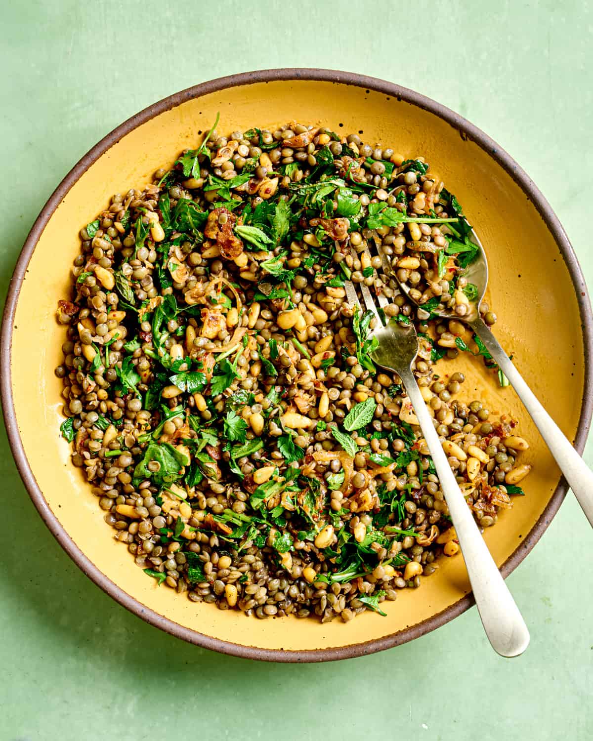 Spoon and fork in lentil salad in a yellow bowl on green table.