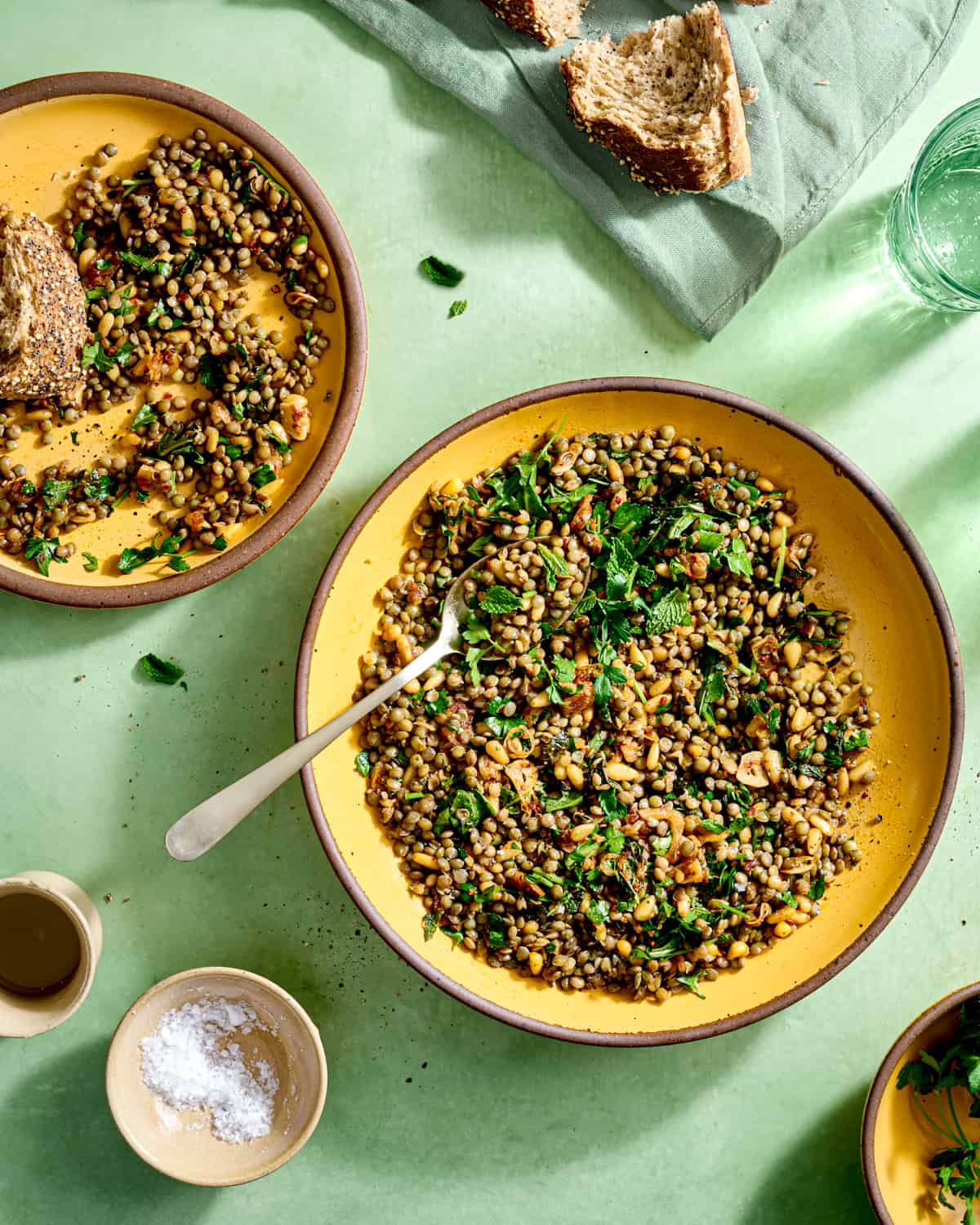 Overhead view of a bowl and a plate of lentil salad and a chunk of bread on a green table.