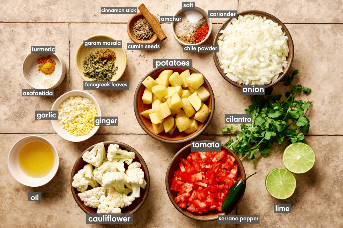 ingredients for Aloo Gobi recipe laid out on light brown tiled surface with ingredients labeled. 