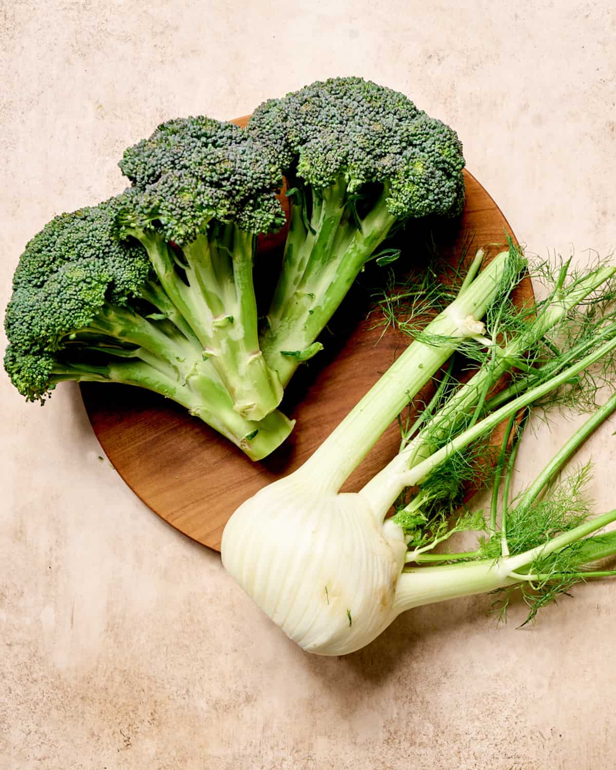 Head of broccoli and fennel on a wooden plate on a light pink surface.