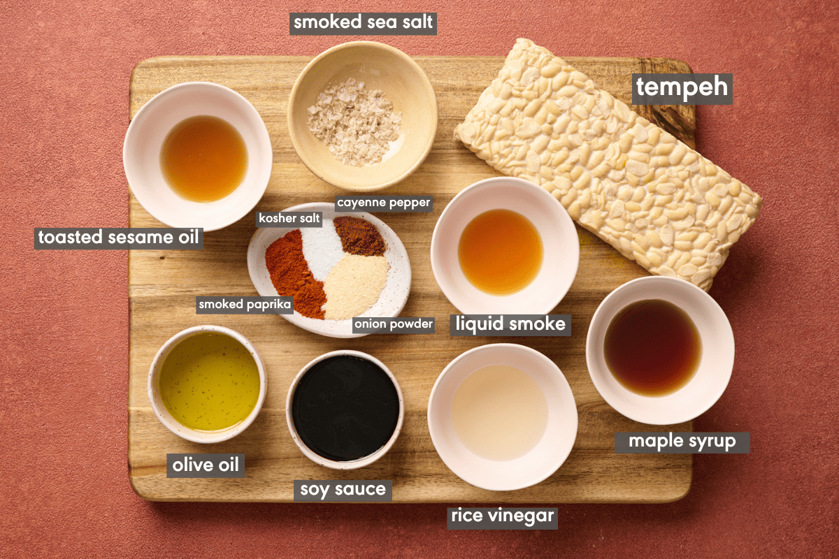 Ingredients for tempeh bacon laid out in bowls and labeled on a wooden cutting board.