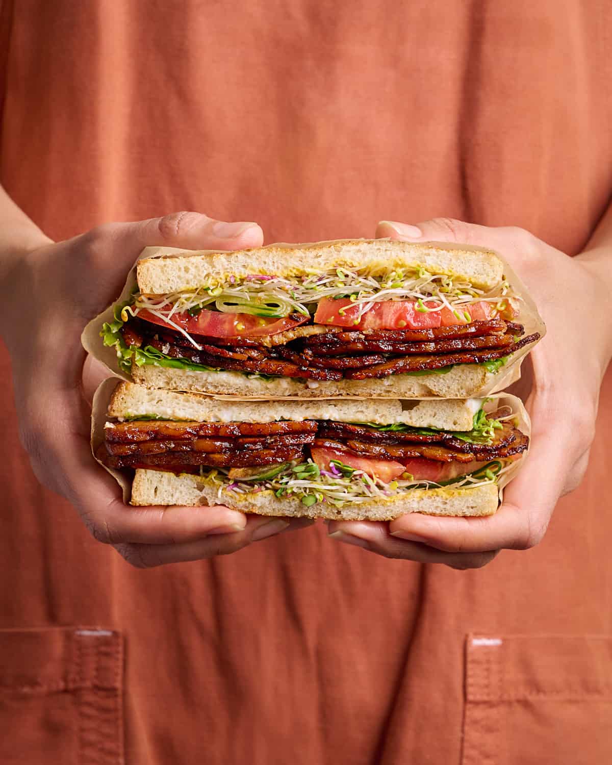 Woman in red apron holding a tempeh bacon vegan BLT sandwich cut in half.