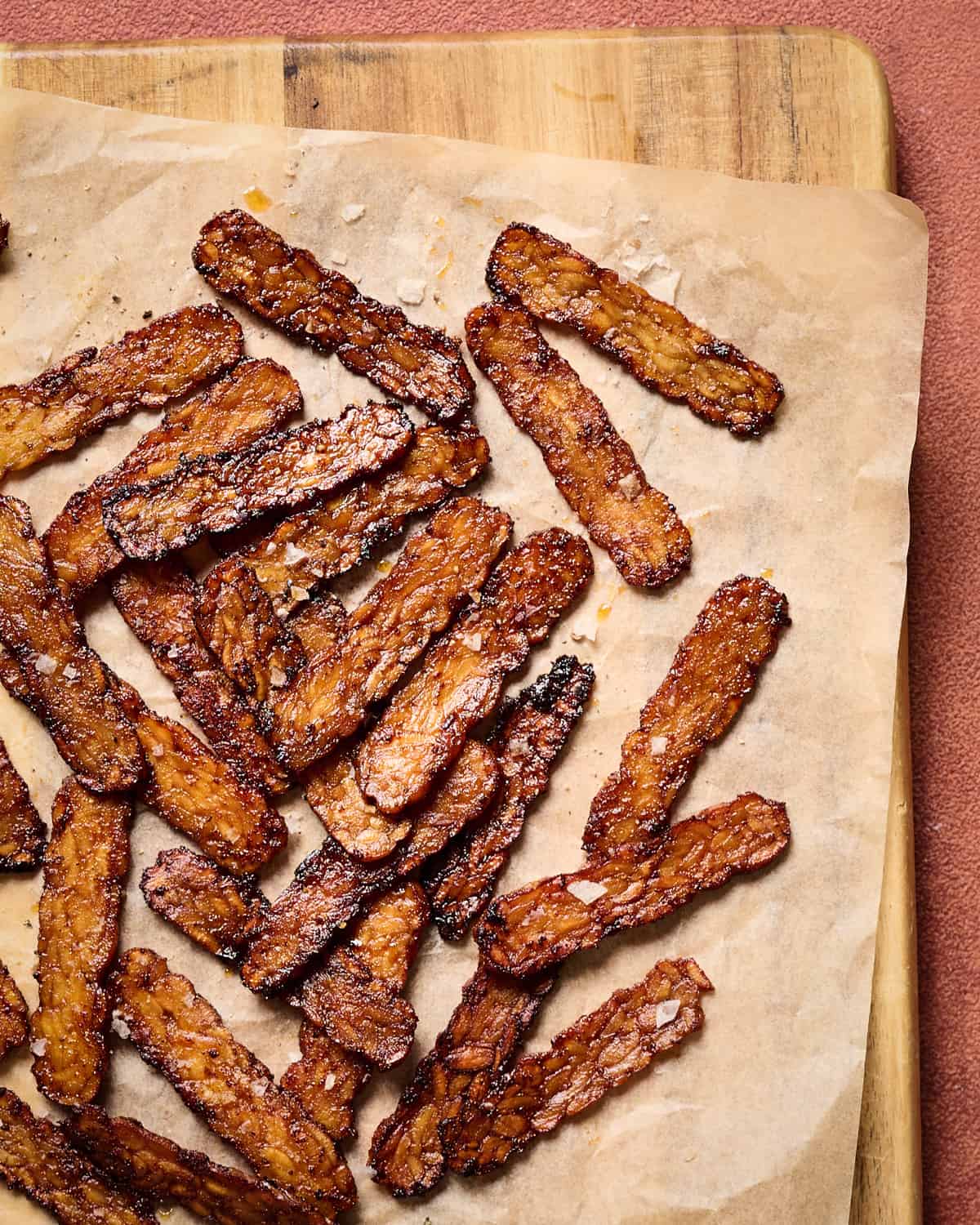 Crispy browned tempeh bacon slices on a piece of parchment paper on a wooden cutting board.