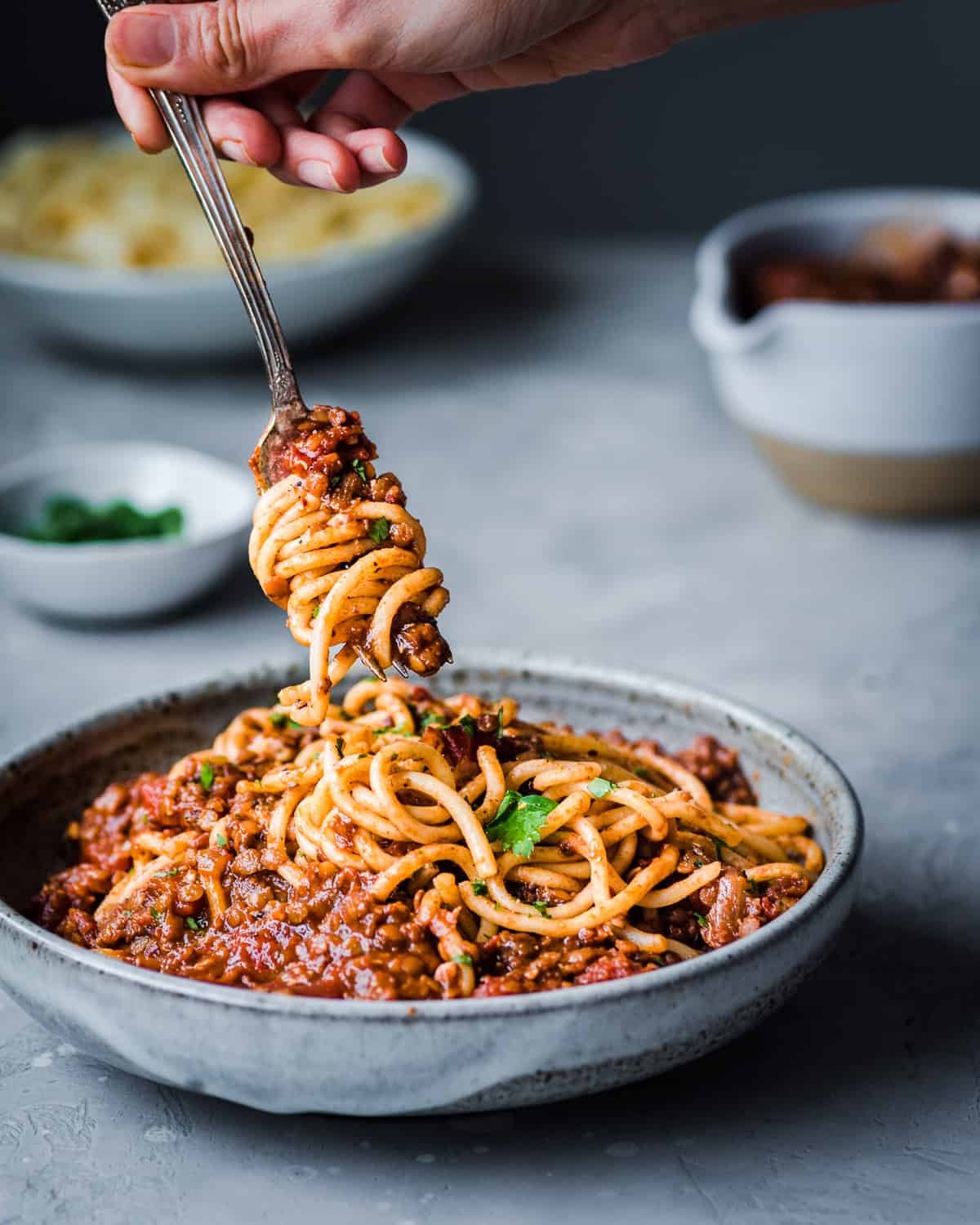 Person twirling lentil bolognese pasta around fork in a bowl on a grey table.