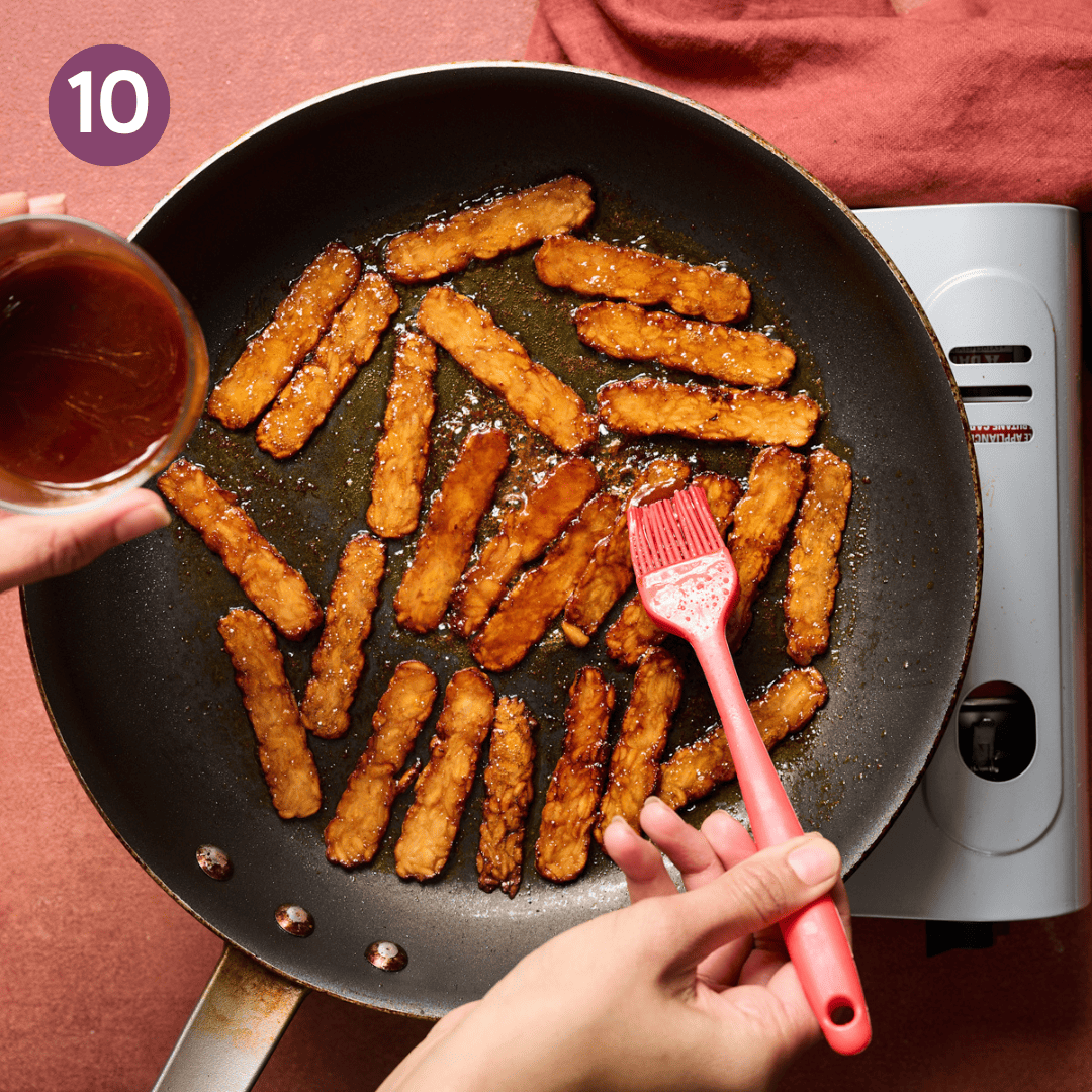 Person brushing even more reserved marinade on the dark colored and almost finished tempeh bacon slices in the pan.