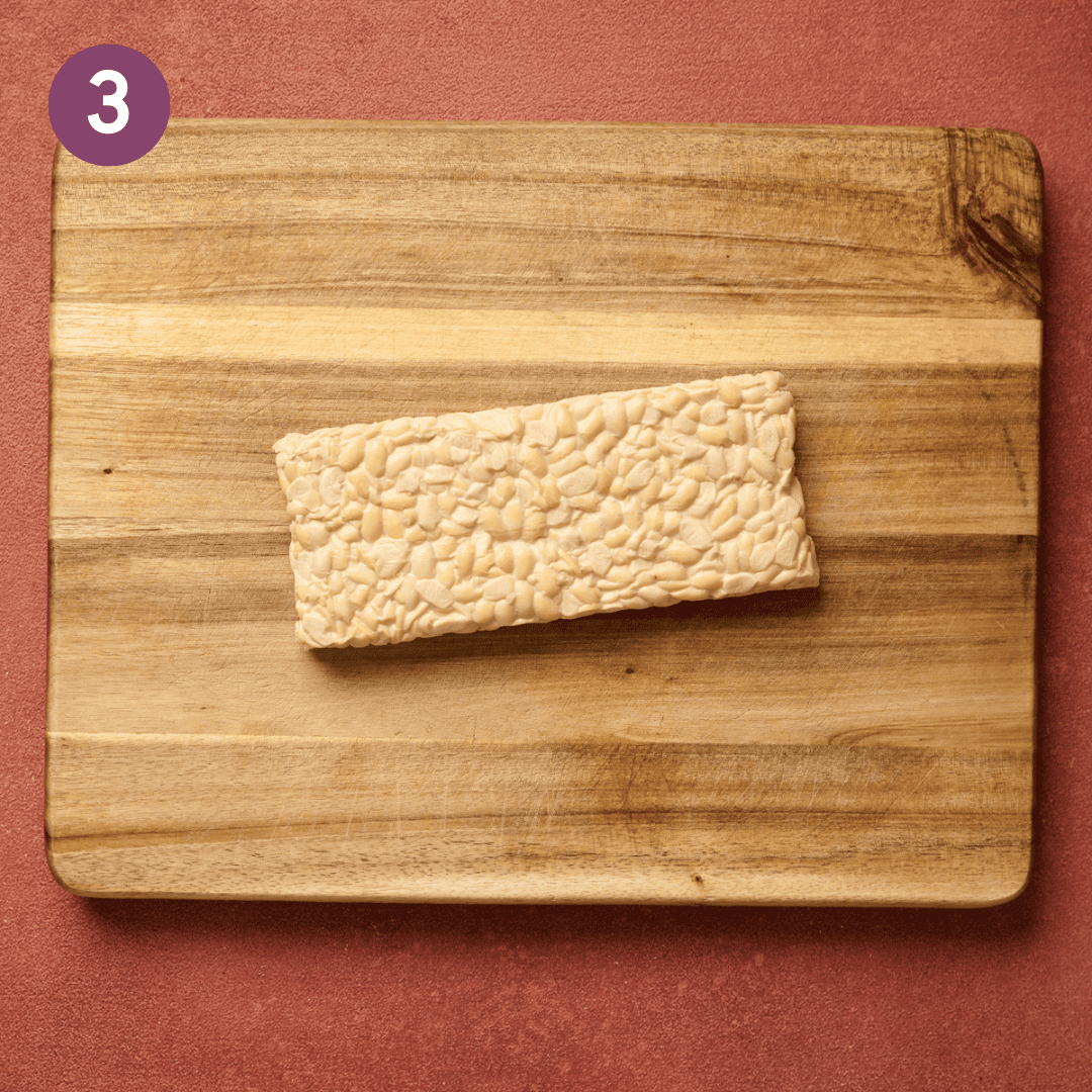Block of tempeh on a wooden cutting board on a red table.