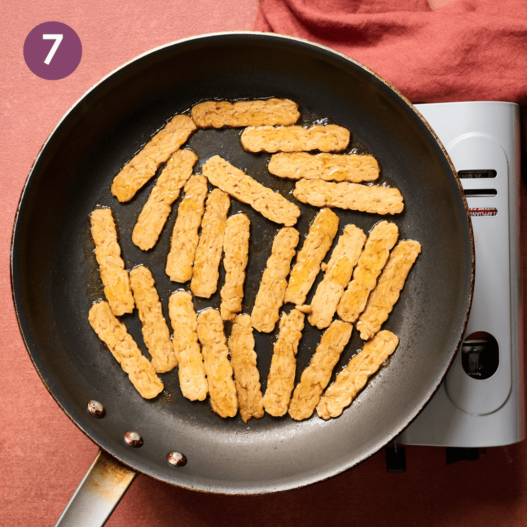 Tempeh bacon strips cooking in a skillet.