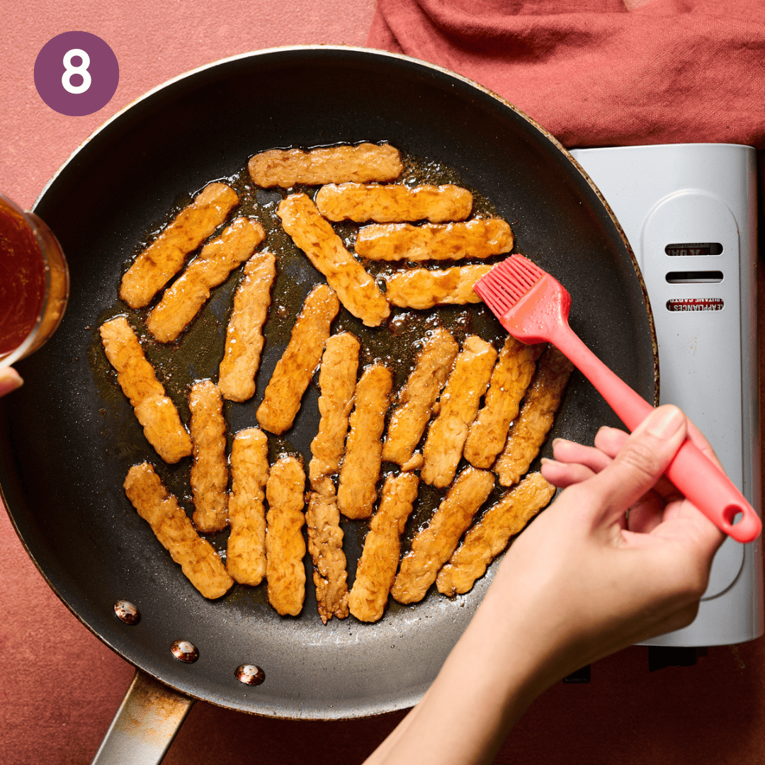 Person brushing reserved marinade onto cooking tempeh bacon slices.